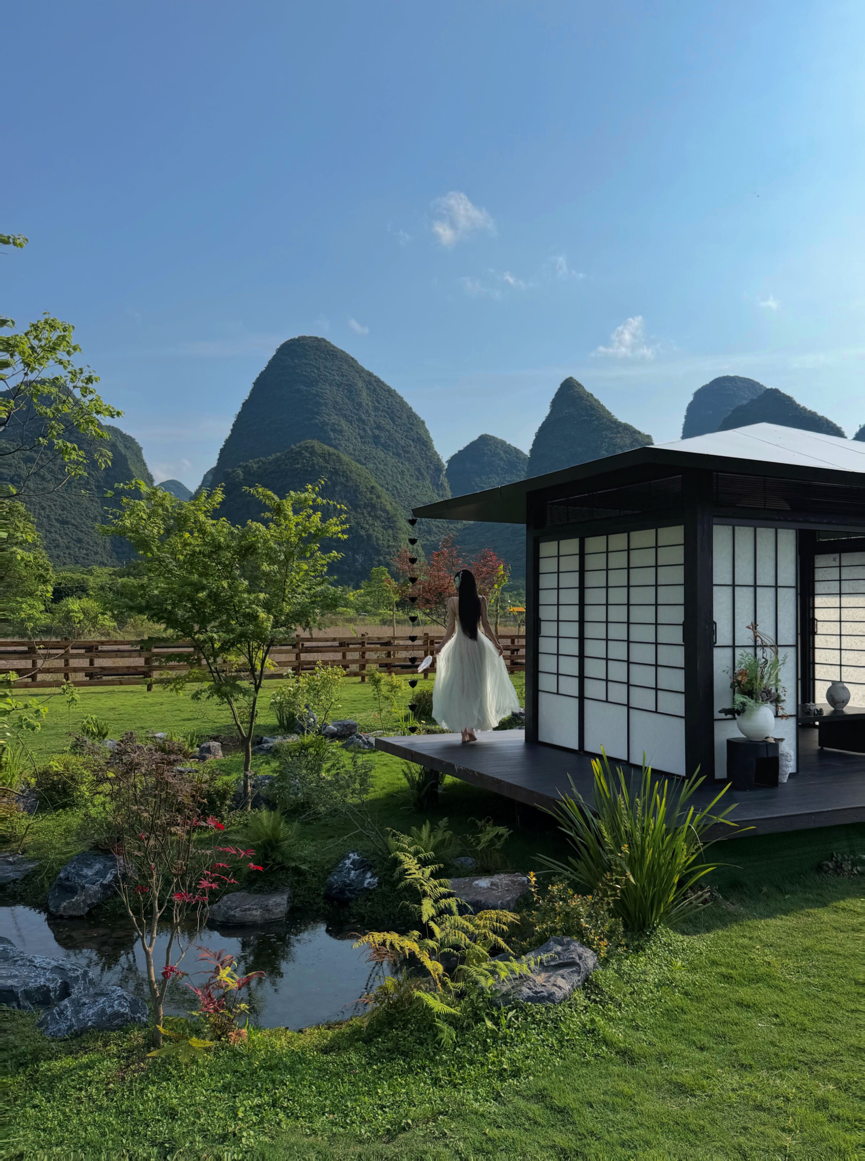 Un edificio tradicional japonés con suelo de tatami, un pequeño jardín con estanque y montañas al fondo.