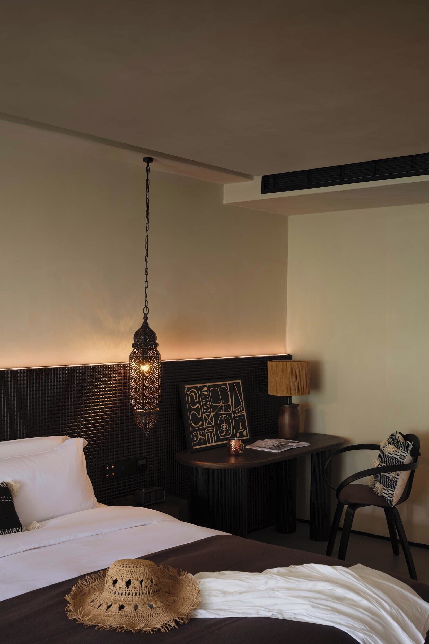 Een hotelkamer met een bed, een hanglamp, een klein tafeltje met een lamp en een stoel.