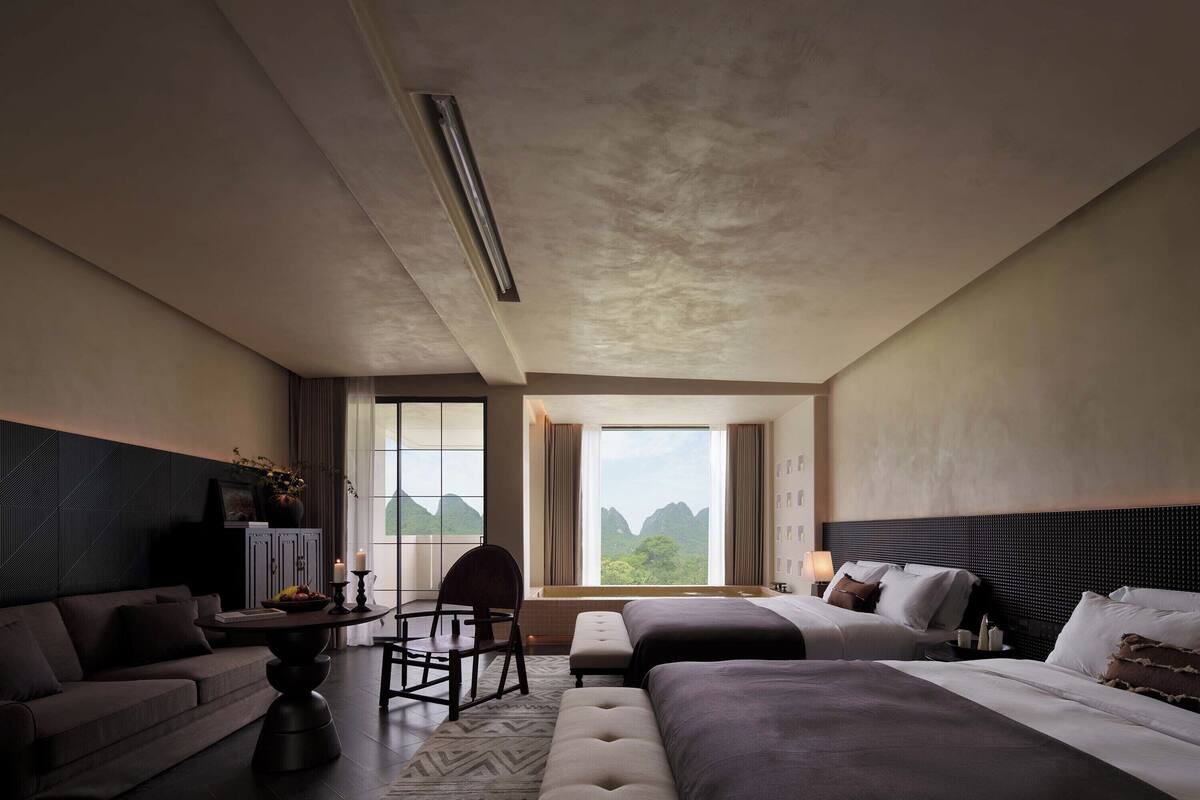 Camera d'albergo con un letto grande, un divano, una poltrona e un tavolino. Si vede una finestra con vista sulle montagne.