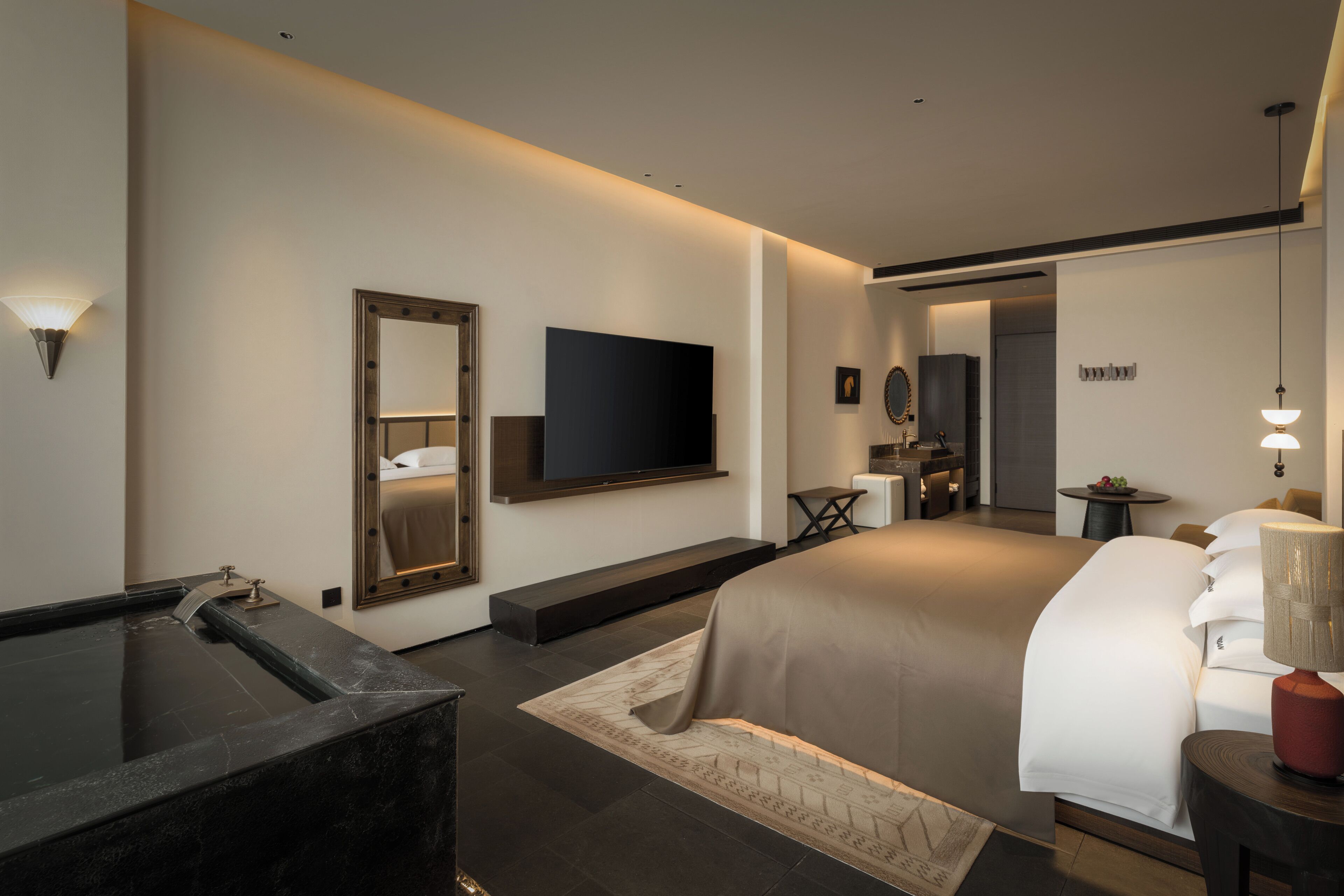 A modern hotel room with a large bed, a flat-screen TV, a mirror, a small table, and a bathroom with a bathtub.