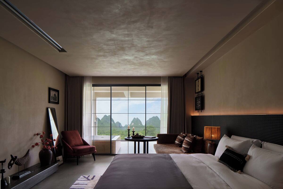 Una camera d'albergo con un letto grande, una zona salotto con divano e poltrone, un tavolino con una lampada, un'ampia finestra vista montagne e uno specchio a parete.