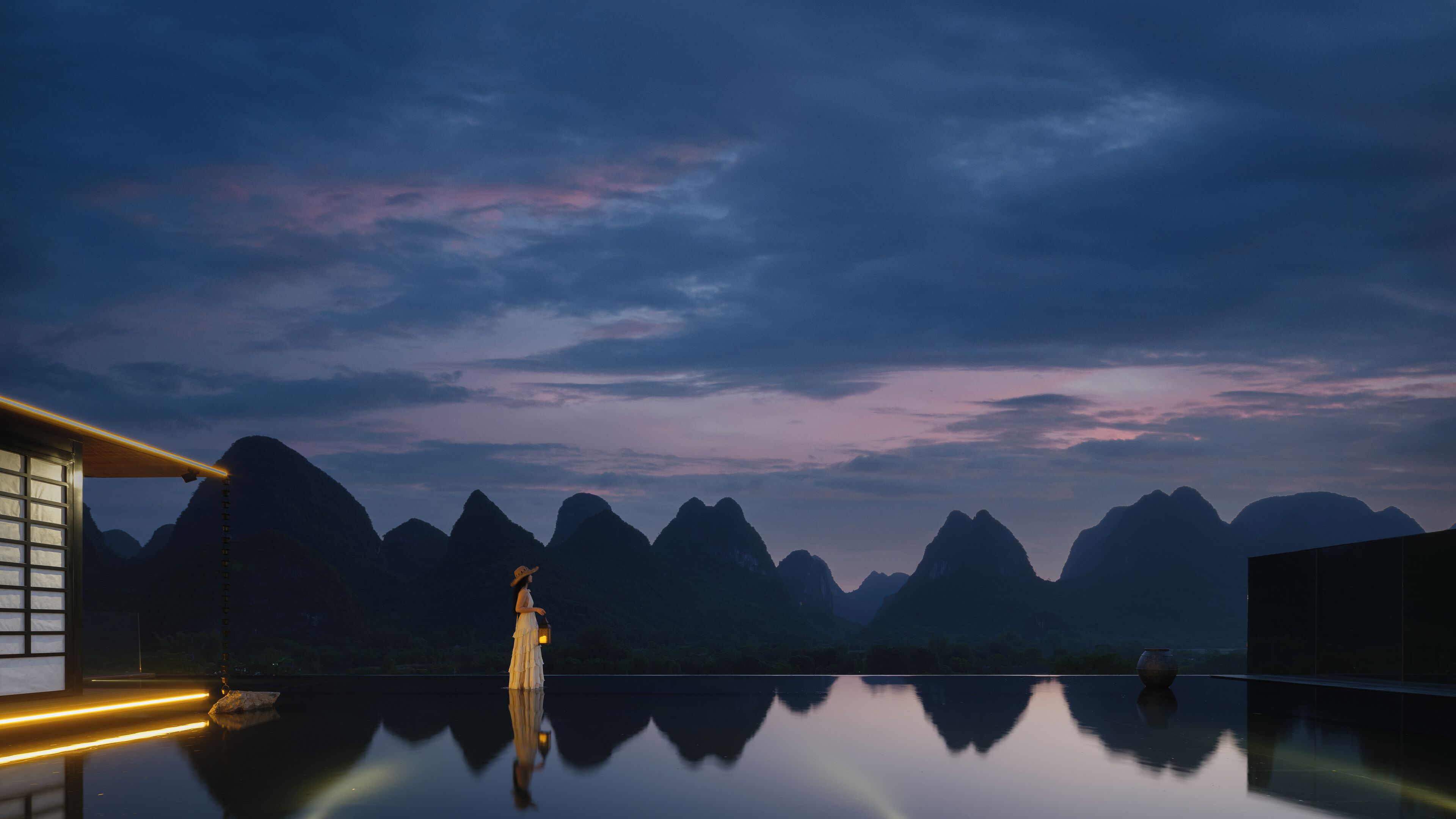 A serene lakeside view with mountain silhouettes and a person holding a lantern.