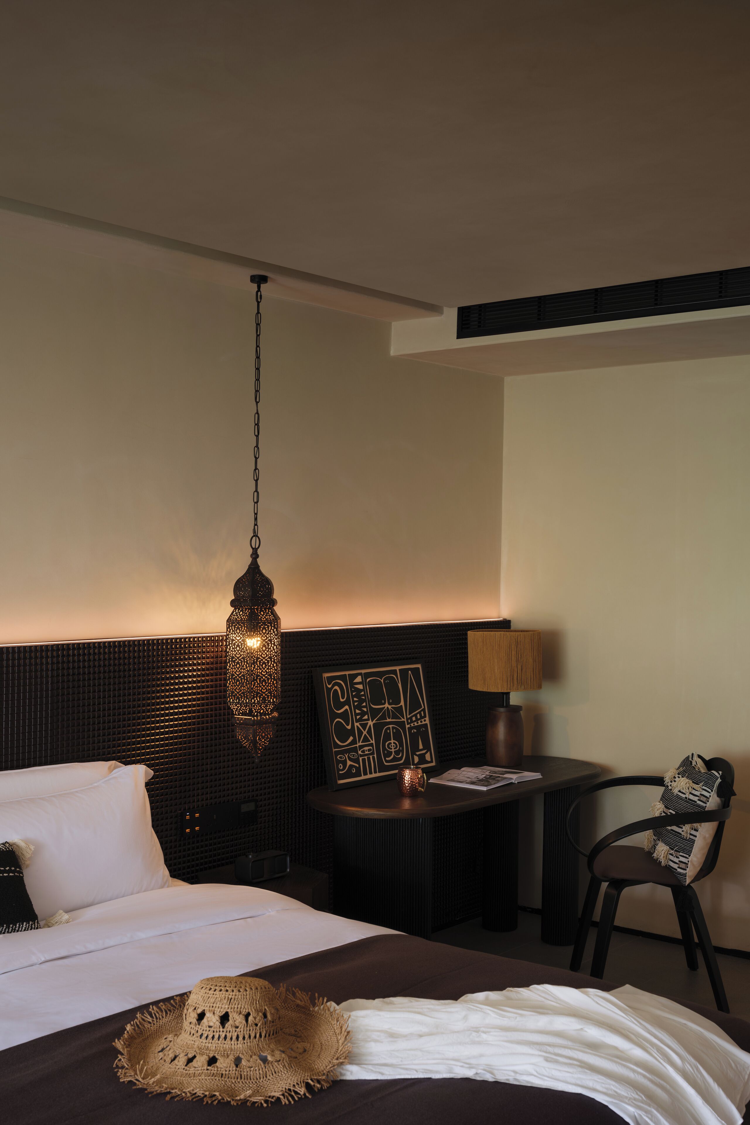A hotel room with a bed, a hanging light fixture, a small table with a lamp, and a chair.