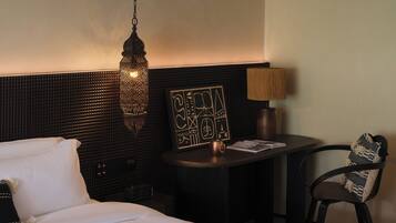 A hotel room with a bed, a hanging light fixture, a small table with a lamp, and a chair.