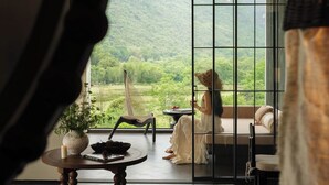 A room with a large window overlooking a lush landscape, a hammock chair, a small table with a candle, and a bed with red cushions. - The Deer Haven Yangshuo - A Luxury Heritage & Modern Karst Mountain Retreat by Yulong River (Guilin)
