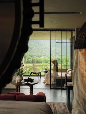 A room with a large window overlooking a lush landscape, a hammock chair, a small table with a candle, and a bed with red cushions. - The Deer Haven Yangshuo - A Luxury Heritage & Modern Karst Mountain Retreat by Yulong River (Guilin)