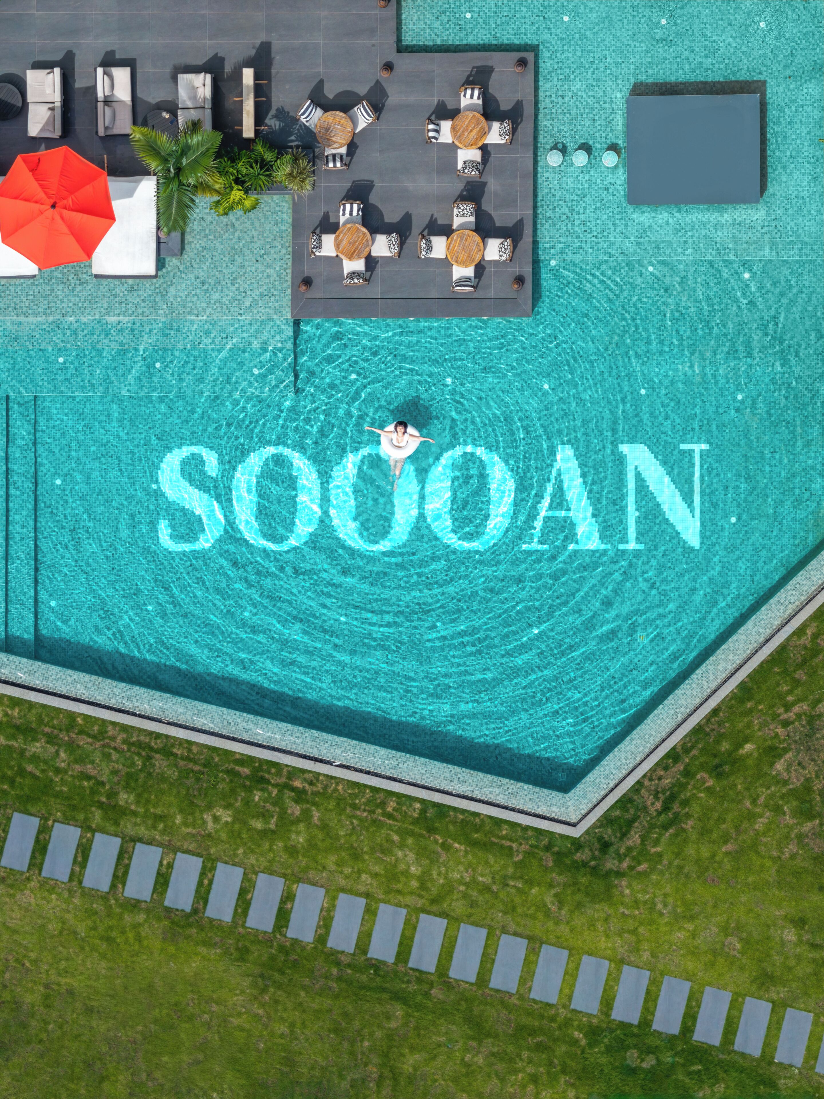 Vista aérea de una piscina con una persona flotando en el centro, rodeada de sillas y mesas de descanso.