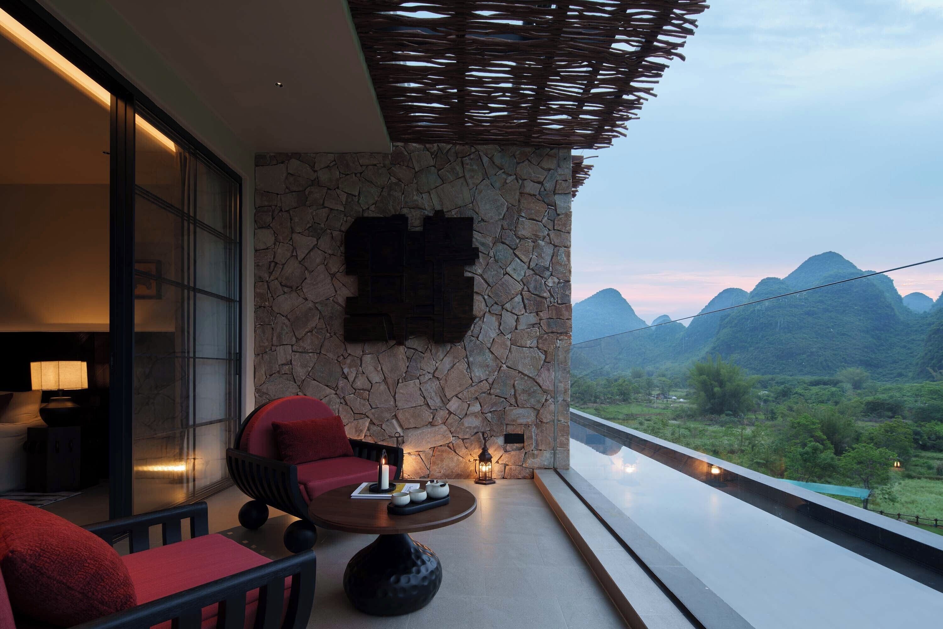 A balcony with a stone wall, a red chair, a round table with candles, and a view of mountains.