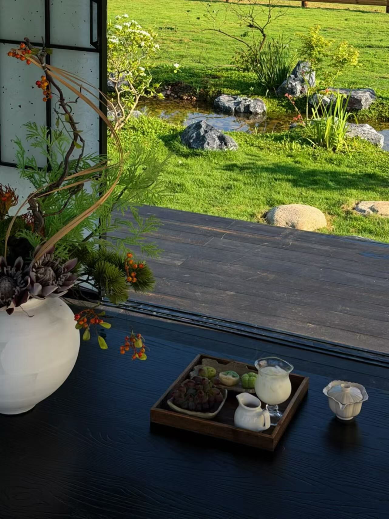 A wooden deck with a tray of food items, a white vase with plants, and a view of a well-maintained garden with rocks and grass.