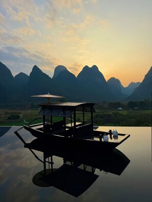 A wooden structure with a roof and a table, surrounded by a reflective water surface, set against a mountainous landscape during sunset. - PastForward Deer Luxury Hidden Retreat at Yulong River  (Guilin)