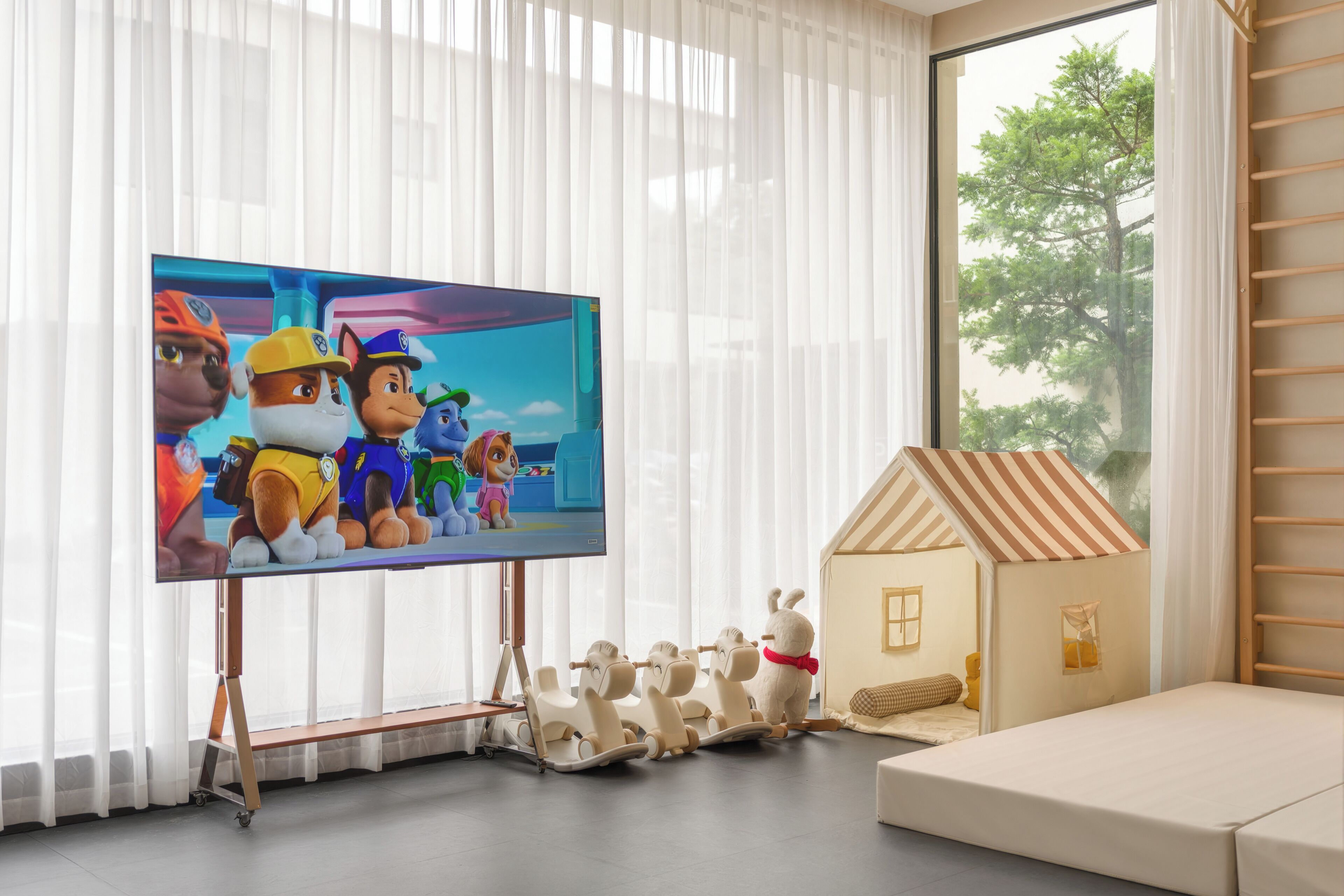 A living room with a flat-screen TV, a wooden playhouse, and a window with sheer curtains.