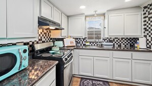 A kitchen with white cabinets, a black and white checkered backsplash, a turquoise microwave, a stove, and a window with blinds. - PRIVATE 2BR GUESTHOUSE: Food Hub & Easy Freeway Access in International District (Houston)