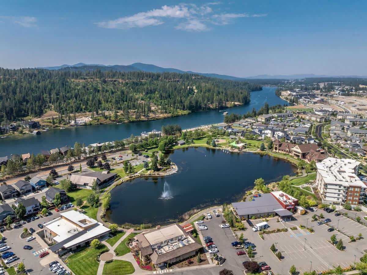 An aerial view of a lakeside community with residential buildings, a central park with a fountain, and a river.