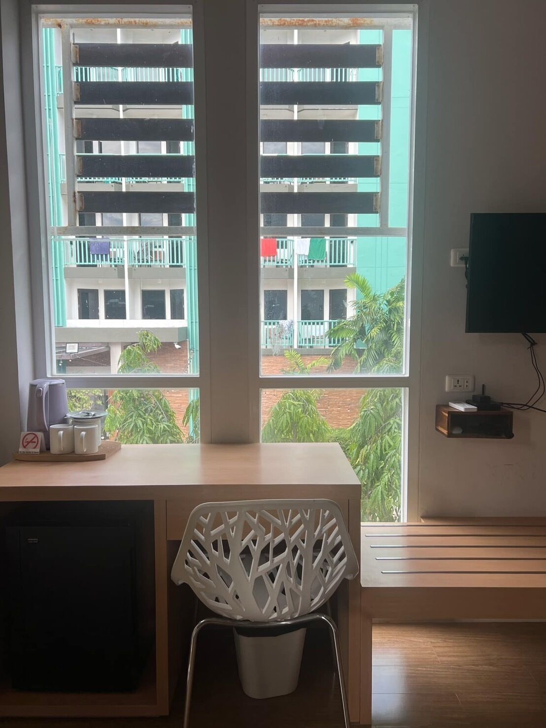 A room with a desk, a chair, a television mounted on the wall, and a window with a view of a building and greenery outside.