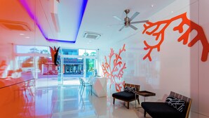 Eine moderne Lobby mit Glastrennwand, einer Rezeption, einer Sitzecke mit Sofa und Sesseln sowie einem Deckenventilator.
