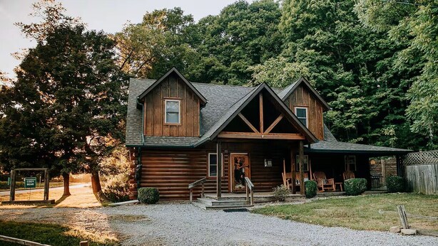 A rustic log cabin with a porch, a gravel driveway, and a forested backdrop. - Cozy Bear Cabin - Log Cabin Soul, Nashville Heart (Nashville)