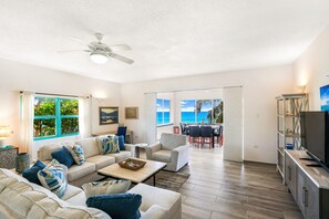 A spacious living room with a large window, a ceiling fan, a dining area, and a TV on a cabinet. - Oceanfront 3BR Condo, Pool, Direct Beach Access (Cole Bay)