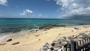 A beach with clear turquoise water, a sandy shore, and a rocky breakwater. - Oceanfront 3BR Condo, Pool, Direct Beach Access (Cole Bay)