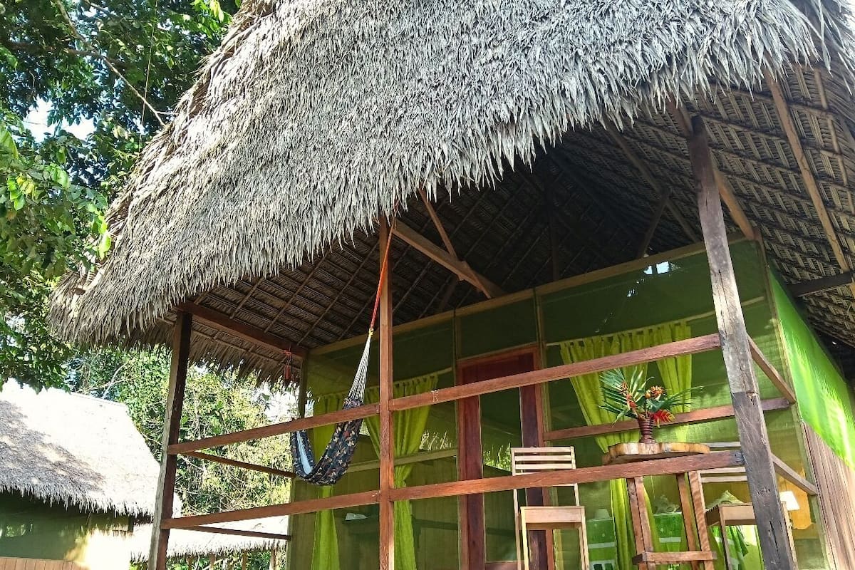 Una cabaña con techo de paja, estructura de madera, una hamaca y una ventana con cortinas.