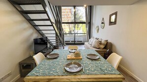 A dining area with a wooden table set for four, a staircase, and a sofa in the background. - Rent Up - Luminoso Duplex en la Boca (Buenos Aires)
