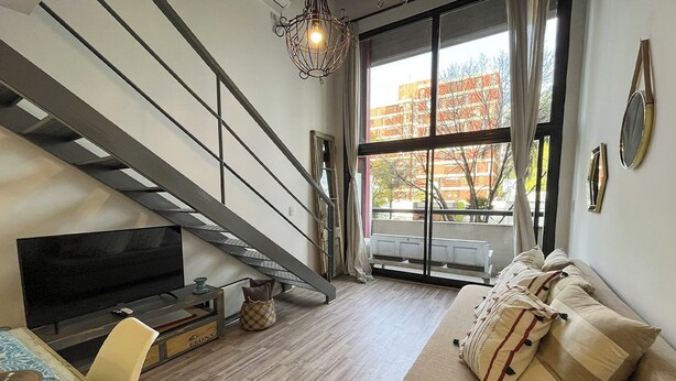 A modern living room with a staircase, a flat-screen TV, a sofa with patterned cushions, and a large window with a view of buildings. - Rent Up - Luminoso Duplex en la Boca (Buenos Aires)