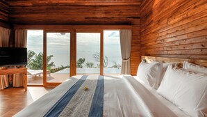 A wooden cabin bedroom with a large bed, a TV on a wooden stand, and a view of the outdoors through large sliding doors. - Seraya Cove Estate by Manara (Karangasem)