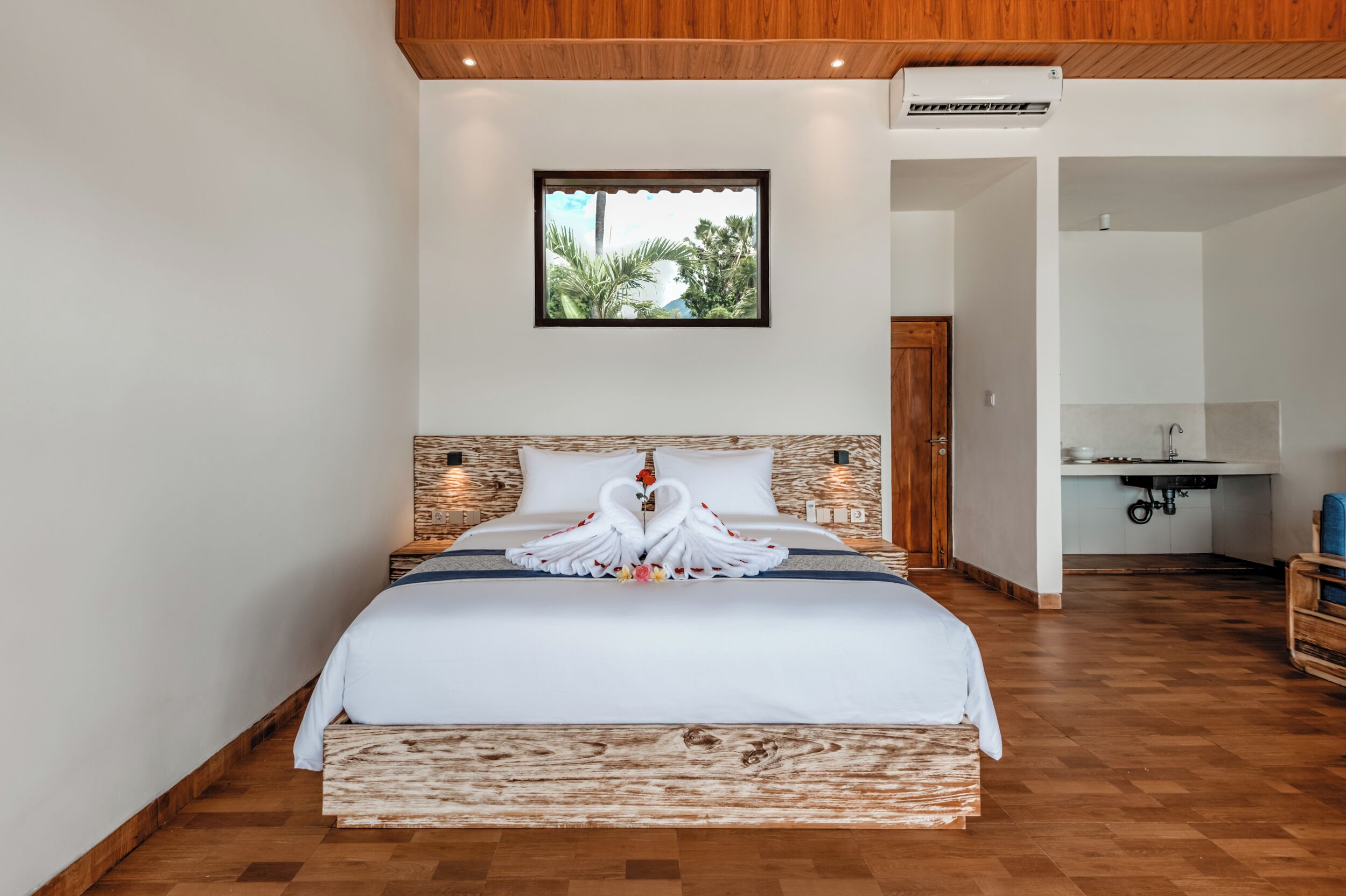 A hotel room with a bed, a wooden headboard, a window with a view of greenery, and a kitchenette area.