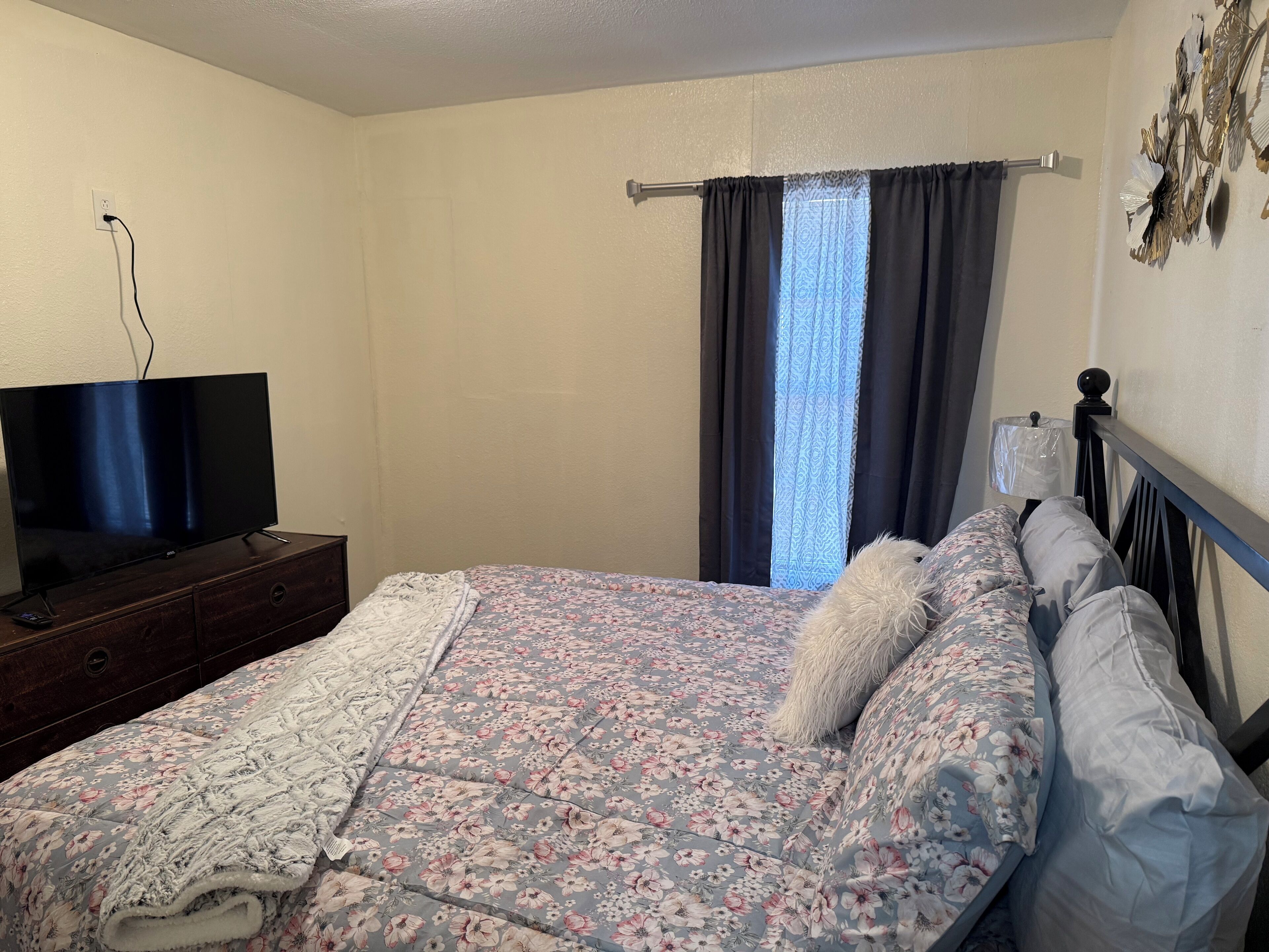 A bedroom with a bed, a TV on a dresser, a window with curtains, and a wall-mounted lamp.