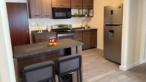 A modern kitchen with wooden cabinets, a stainless steel refrigerator, a microwave, and a sink. There is a small island with two chairs and a red toy character. - REMODELED Stellar Yellow House w/mini cine, near Plaza Mall & Downtown McAllen (McAllen)