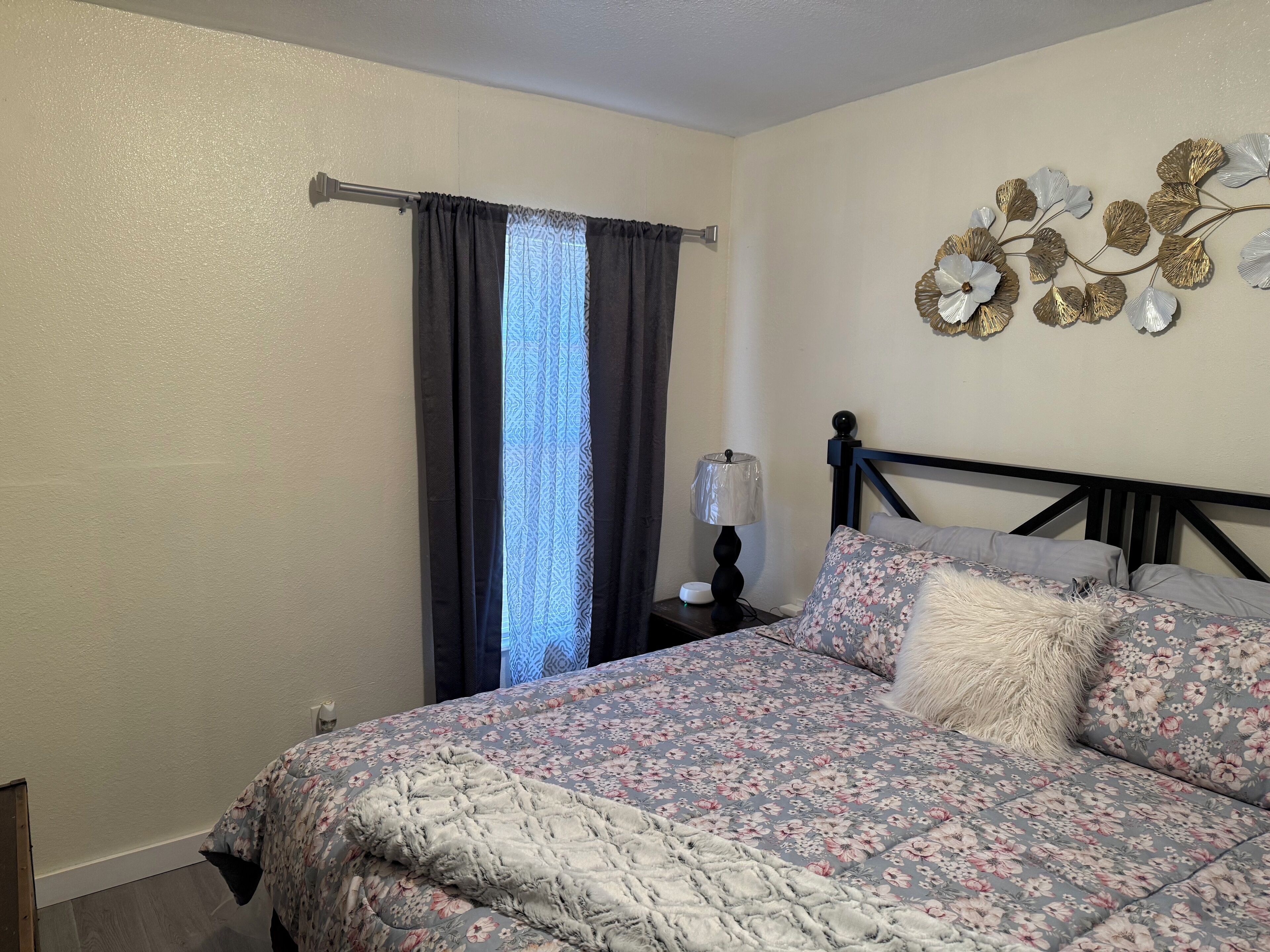 A bedroom with a bed, a nightstand, a lamp, a window with curtains, and a wall decoration.