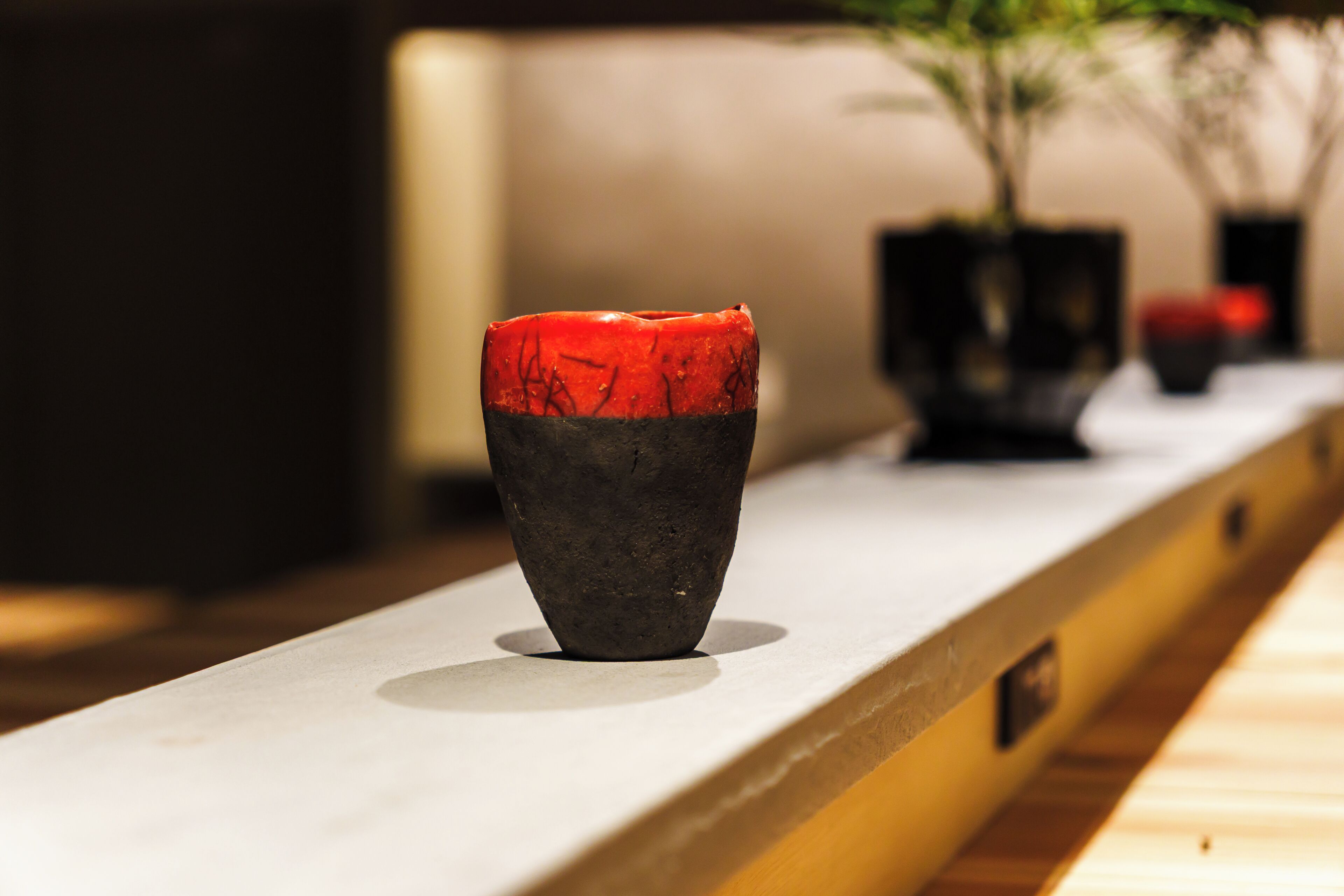A dark-colored vase with a red top on a white surface, with a blurred plant and a black object in the background.