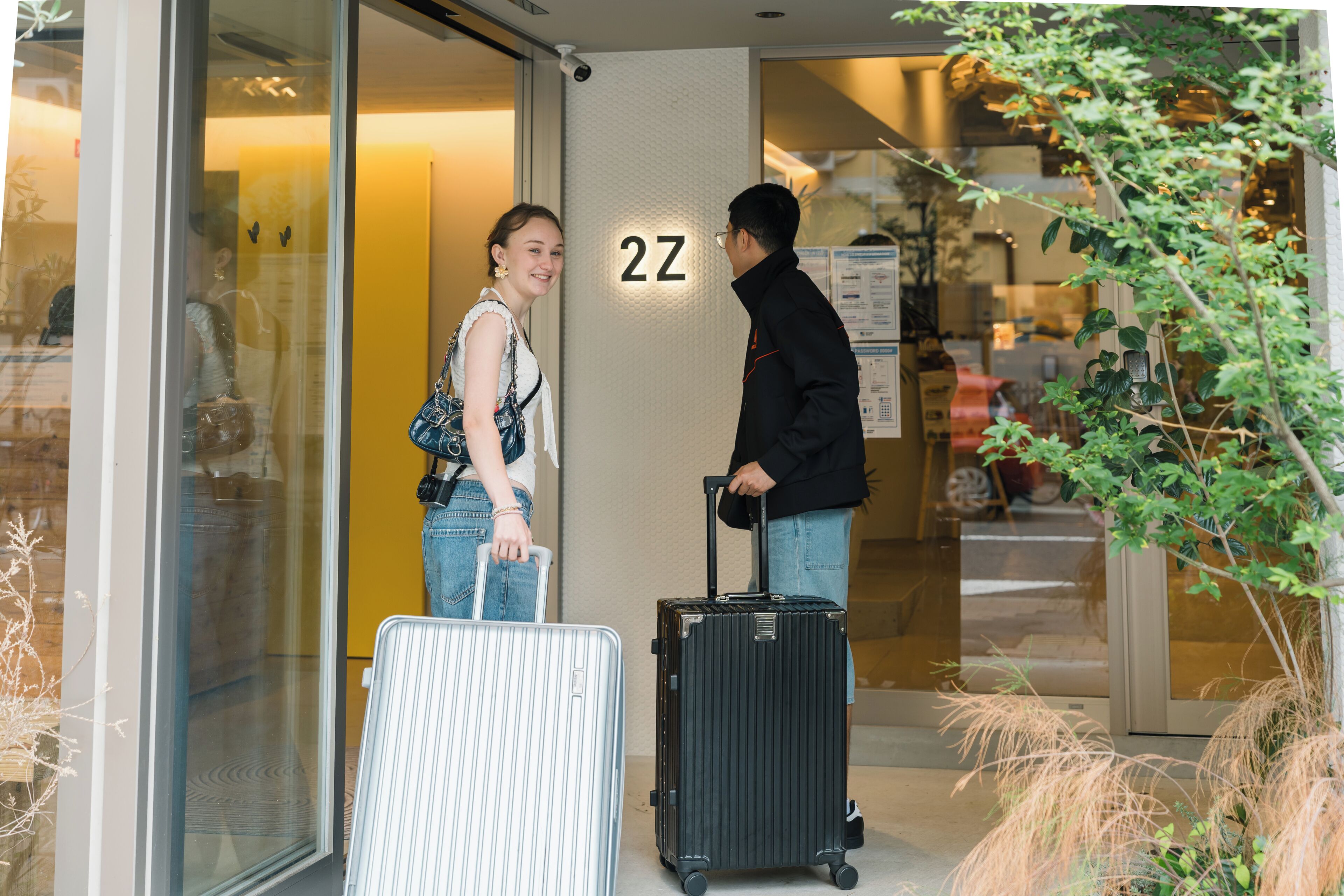 L’entrée d’un hôtel, avec une porte en verre, une enseigne portant le numéro « 2Z », et une personne tenant une valise.