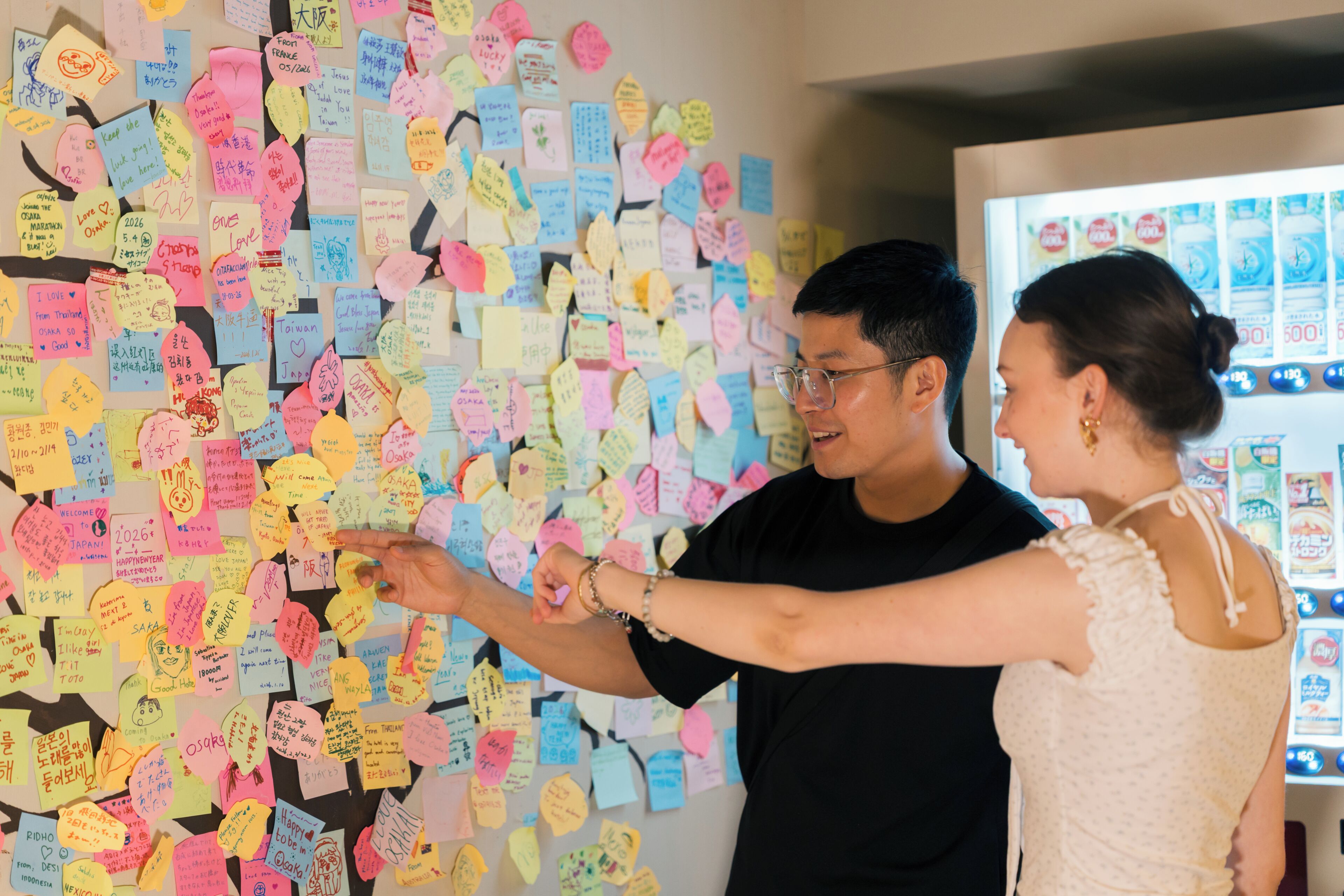 A wall covered with colorful sticky notes, a vending machine, and a person pointing at the notes.