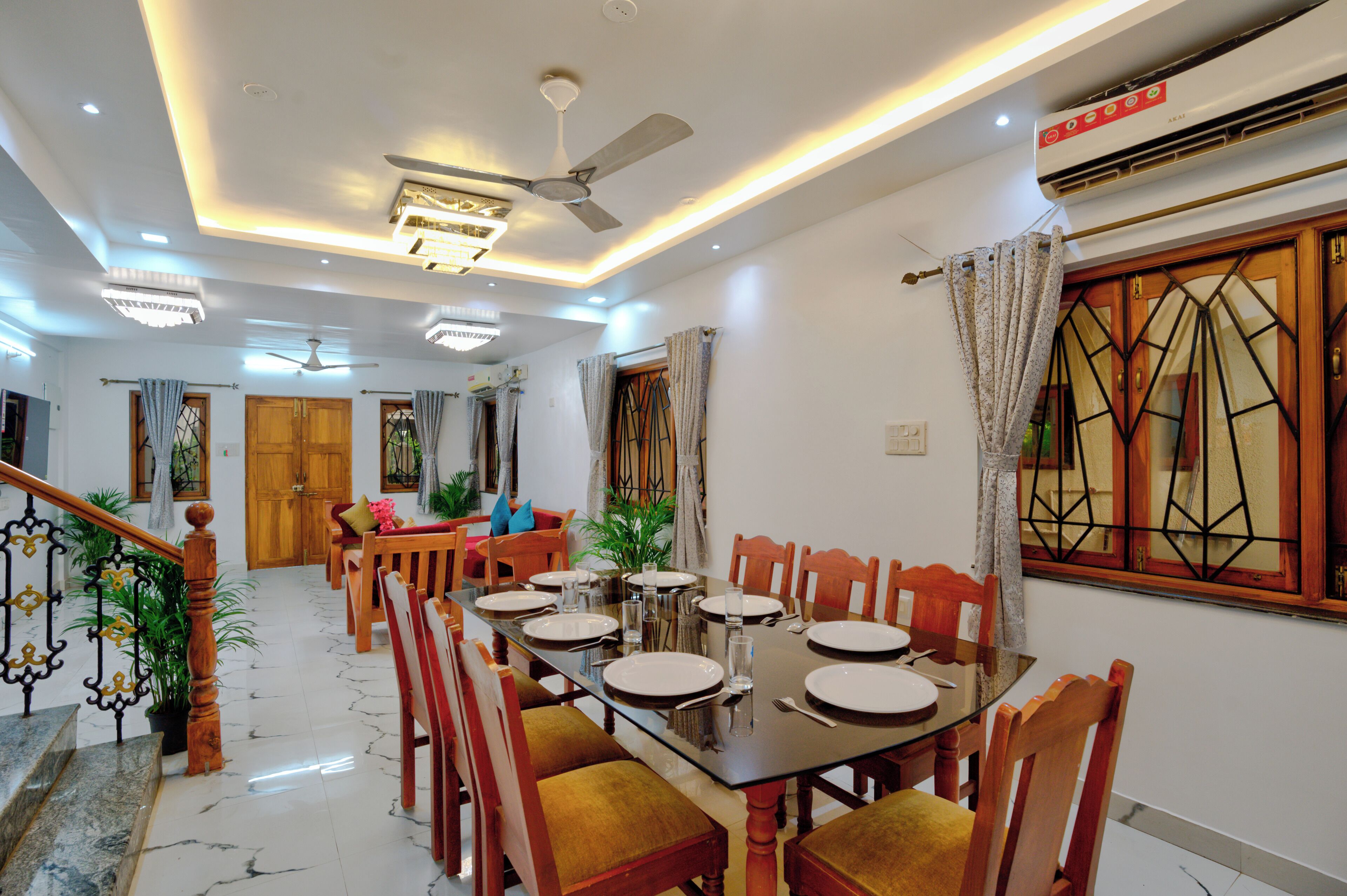 A dining area with a large table set for six, surrounded by wooden chairs. There is a staircase with a railing leading to another room.