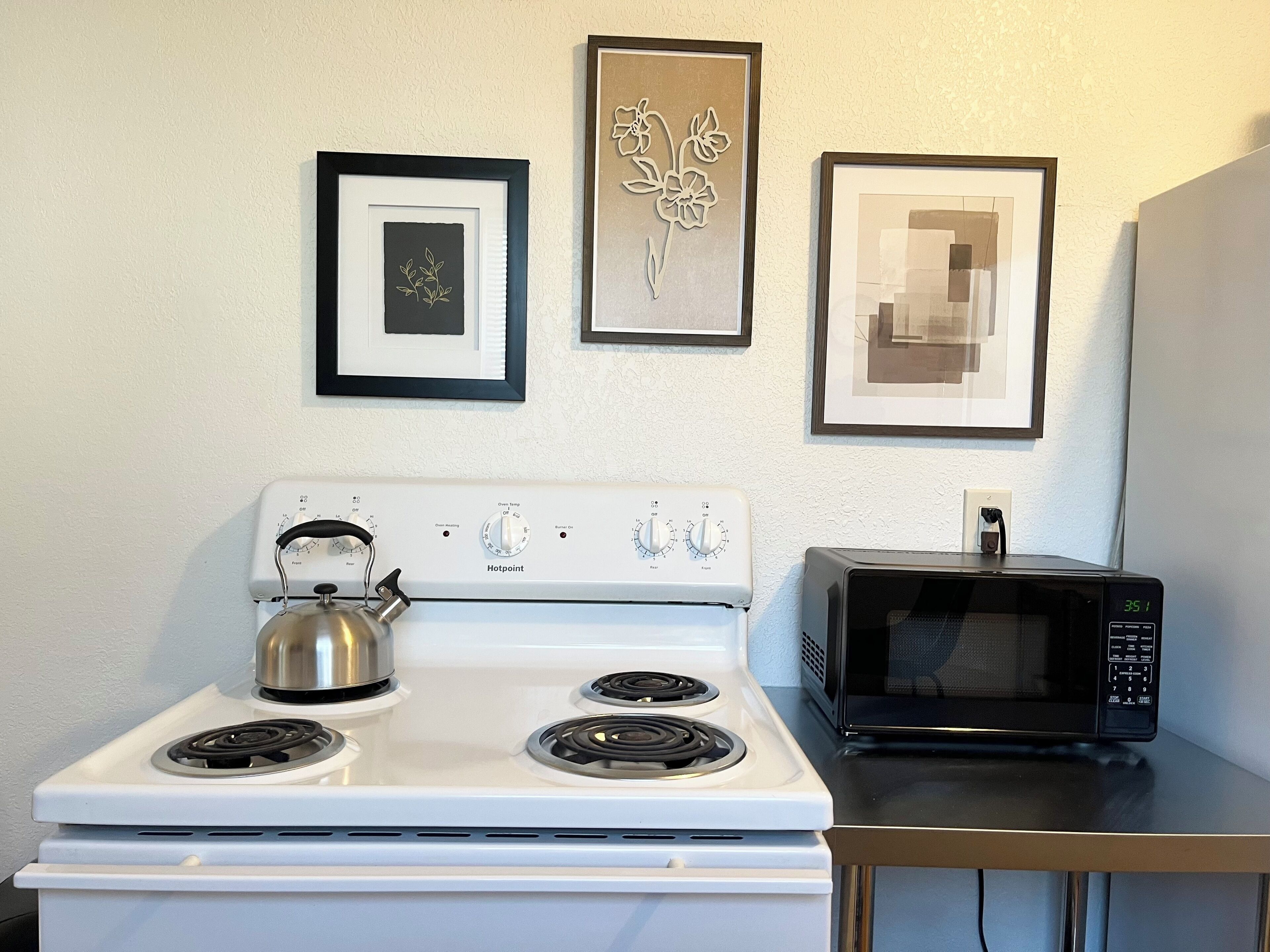 Une cuisine avec un four blanc, une bouilloire et un micro-ondes noir. Trois tableaux encadrés sont accrochés au mur.