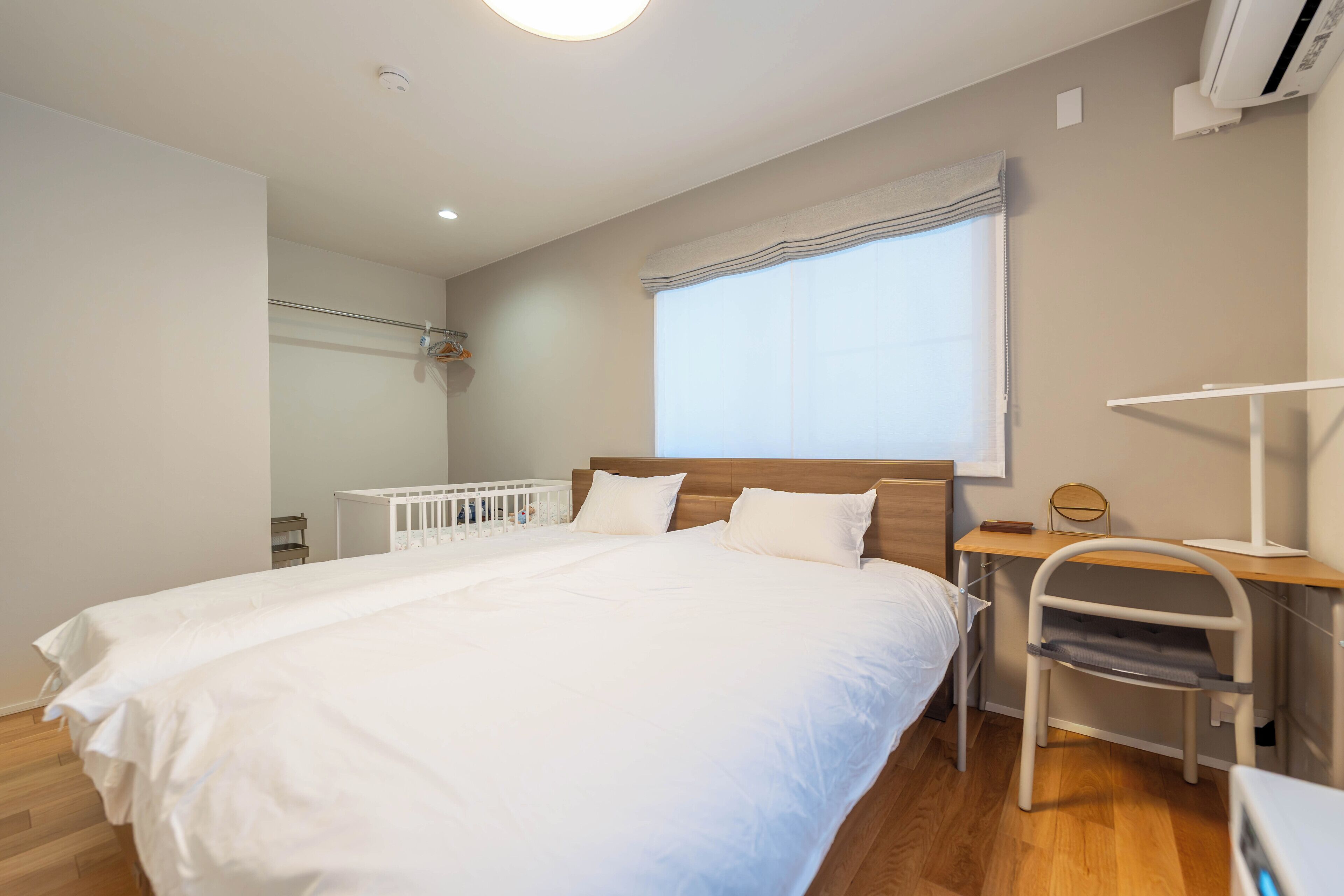 A bedroom with a large bed, a desk with a chair, a window with blinds, and an air conditioning unit.