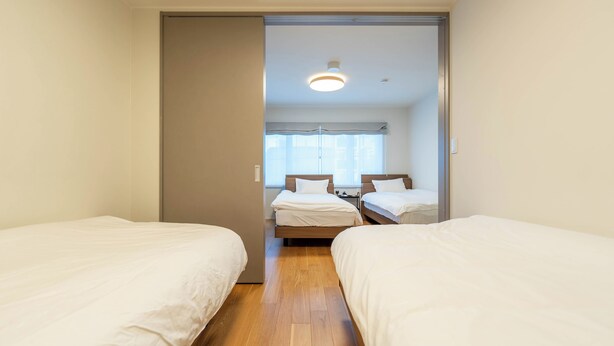 Ein Hotelzimmer mit zwei Einzelbetten, Holzboden, einer Deckenleuchte und einem mit Vorhängen versehenen Fenster.