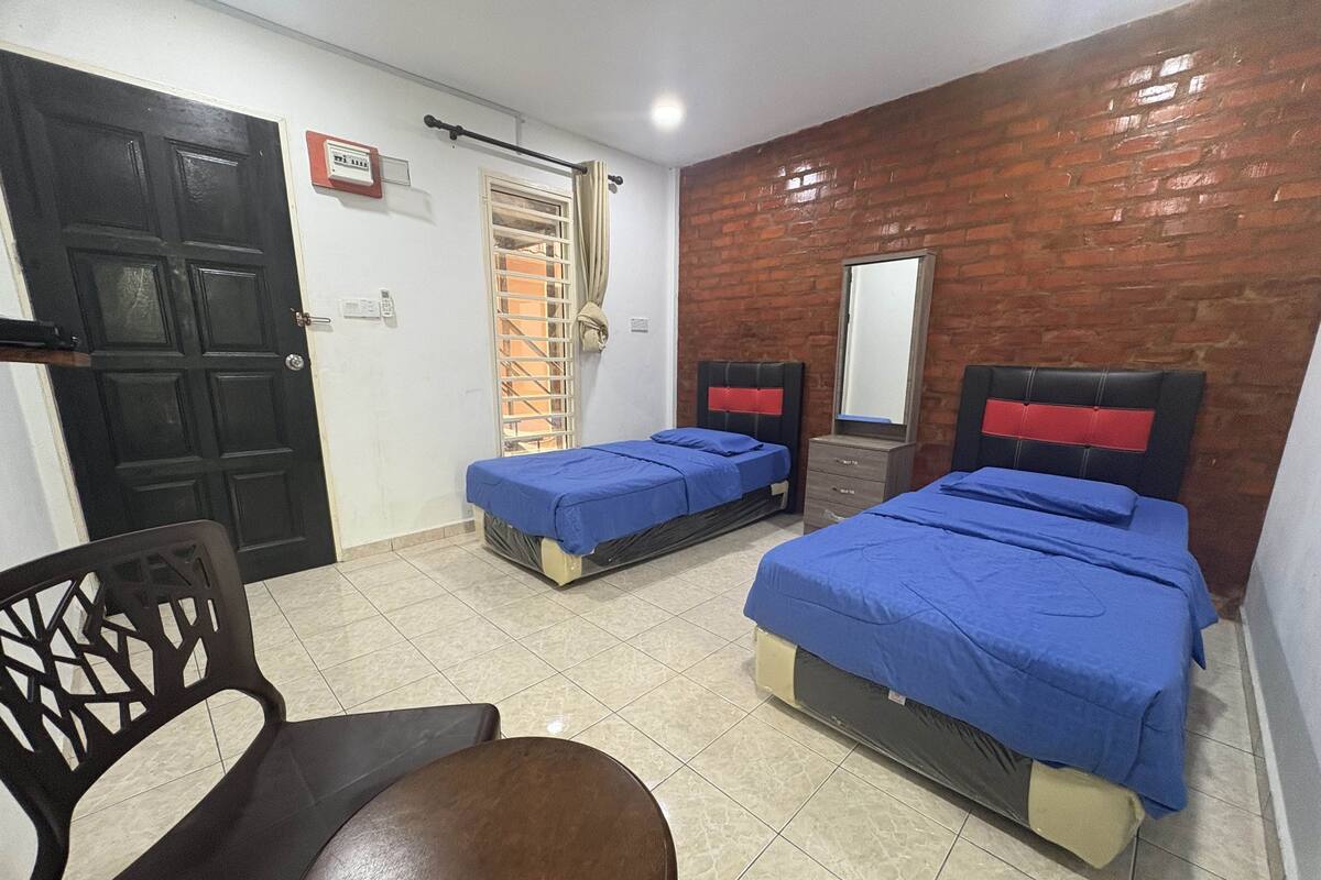 A room with two beds, a chair, a desk, a window with curtains, a door, and a brick wall.