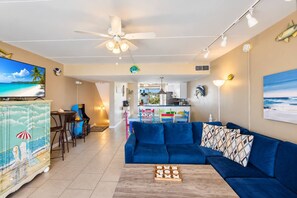A living room with a blue sofa, a coffee table with a game, a TV, and a ceiling fan. - Gulf-Front Villa A-6 Pool Access Sleeps 7 (Saint George Island)