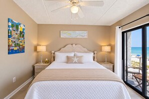 A hotel room with a bed, two bedside tables, a ceiling fan, a window with a beach view, and a map on the wall. - Gulf-Front Villa A-6 Pool Access  Sleeps 7 (Saint George Island)