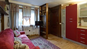Un ambiente compatto con un divano rosso, un tappeto decorato, una televisione e un angolo cottura con mobili rossi.