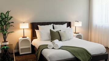 A neatly made bed with white linens and green accents, flanked by two bedside tables with lamps, a potted plant, and a view of a window with sheer curtains.