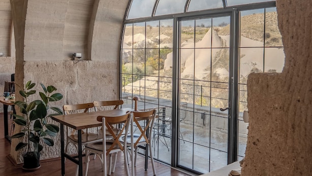 Een ruime binnenruimte met een hoge gewelfde plafond, een houten eettafel met stoelen, een beklede bank en een groot raam met uitzicht op een woestijnlandschap.
