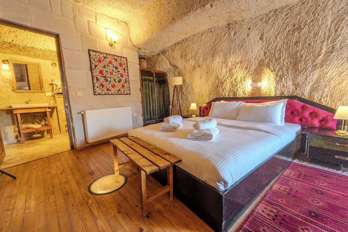 A bedroom with a wooden floor, a bed with white linens, a bench, a rug, and a stone wall.