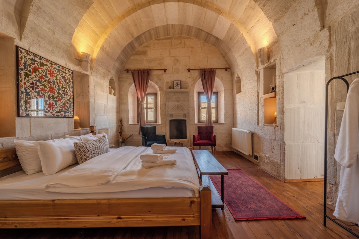 A room with a large bed, wooden furniture, a fireplace, and stone walls.