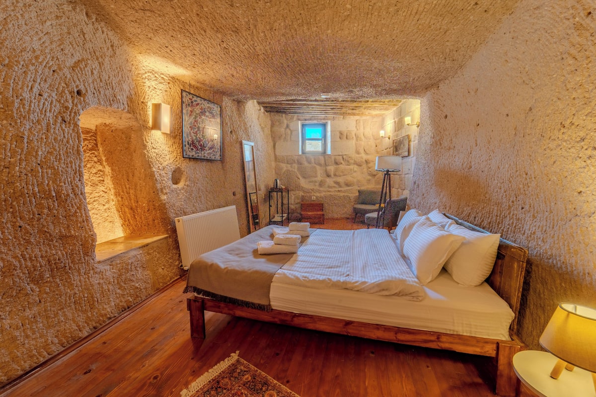 A bedroom with a stone wall, wooden floor, a bed with white linens, a bedside table, a lamp, and a small window.