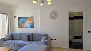 A modern living room with a grey sectional sofa, a painting, a decorative wall clock, and a view into the laundry room. - Brisa Del Mar Beachfront Apartment 116 (Rrashbull)