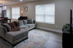 Una sala de estar con un sofá gris, una mesa de centro, un mueble para televisión y un comedor con una mesa y sillas.