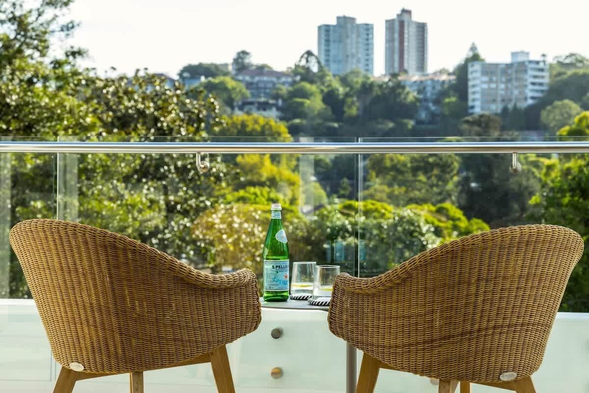 Two wicker chairs with a bottle and glasses on a balcony overlooking a cityscape.
