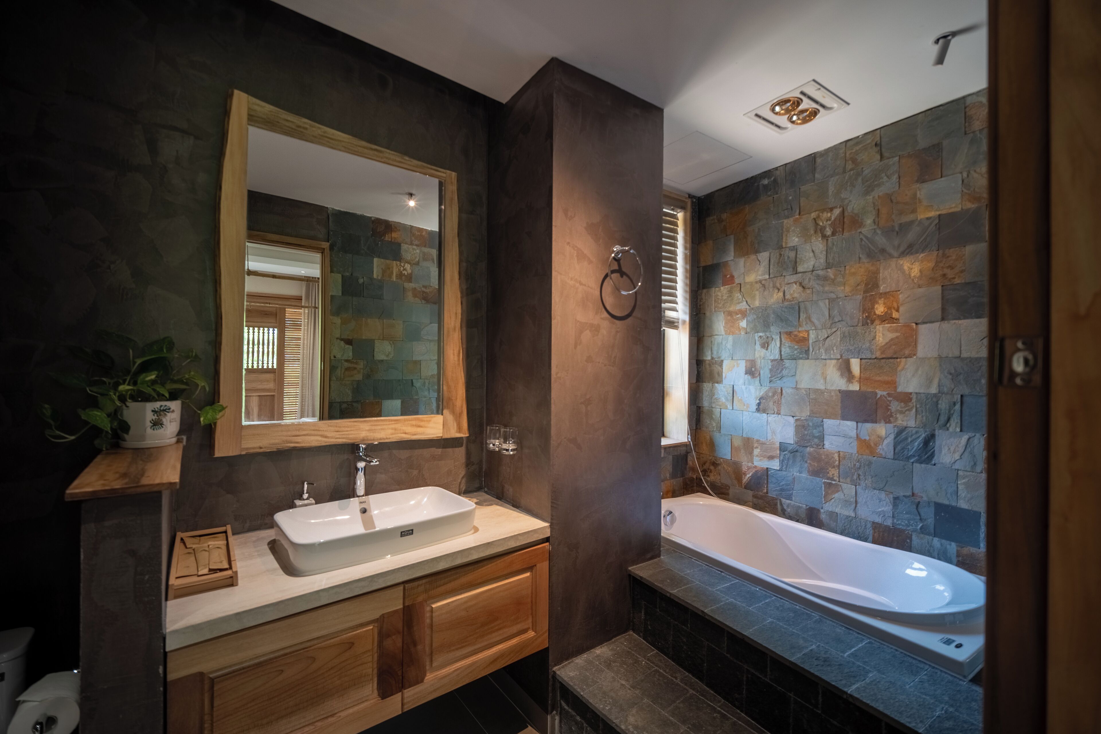 A modern bathroom with a stone mosaic wall, a wooden vanity, a large mirror, and a freestanding bathtub.